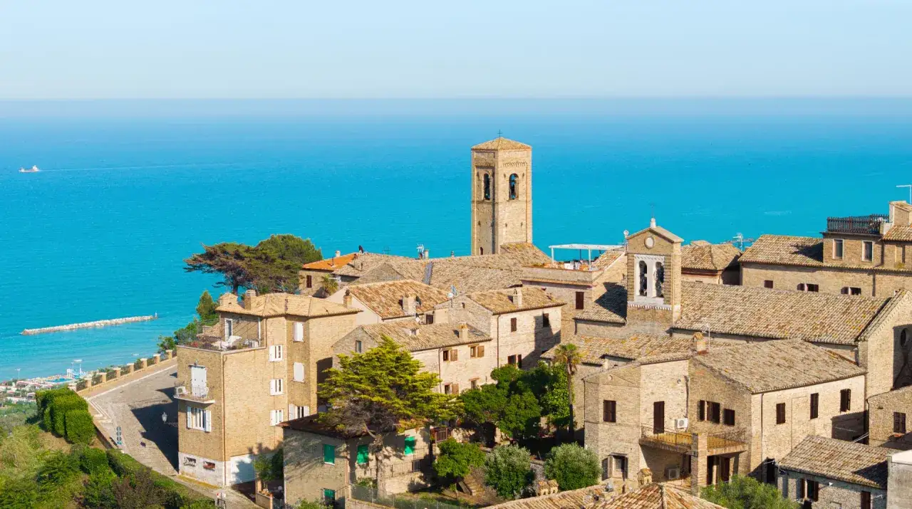 Borgo con torre campanaria affacciato sul mare blu. Case con tetti in cotto e vegetazione rigogliosa.