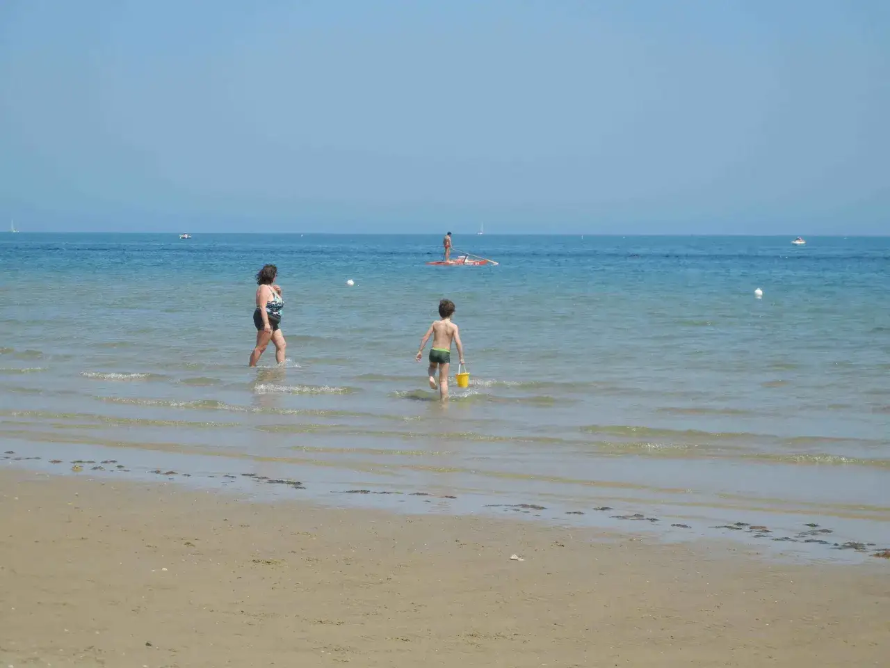 Bambino con secchiello giallo corre verso il mare a Pesaro. Una donna cammina nell'acqua.