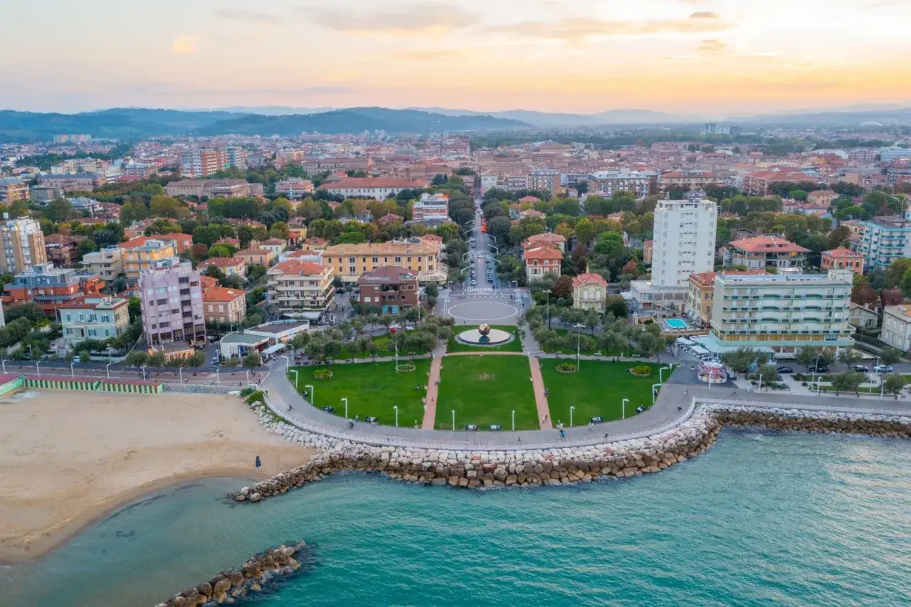 Vista aerea del Pesaro lungomare con la spiaggia, il mare turchese, un parco verde curato e la citt&agrave; che si estende all'orizzonte al tramonto.