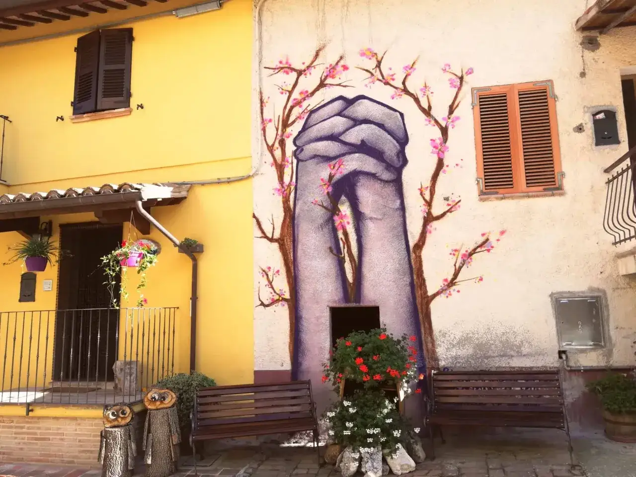 Braccano, paese dei murales: mani giunte e fiori di ciliegio dipinti su un muro, con panchine e decorazioni in legno.