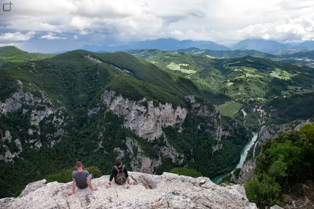 Due escursionisti ammirano la Gola del Furlo, un canyon verdeggiante con un fiume turchese che serpeggia tra montagne imponenti.