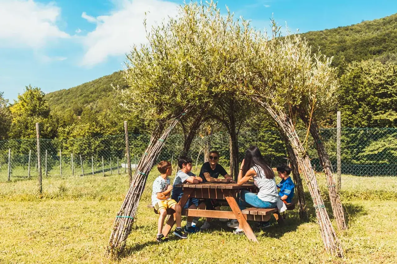 Bambini riuniti attorno a un tavolo da picnic sotto una tettoia naturale nel bosco dei folletti Urbania, immersi nella natura.