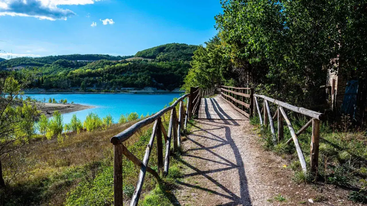 Sentiero in legno con parapetto rustico che costeggia un lago azzurro e colline verdi nella valle di Norcia.