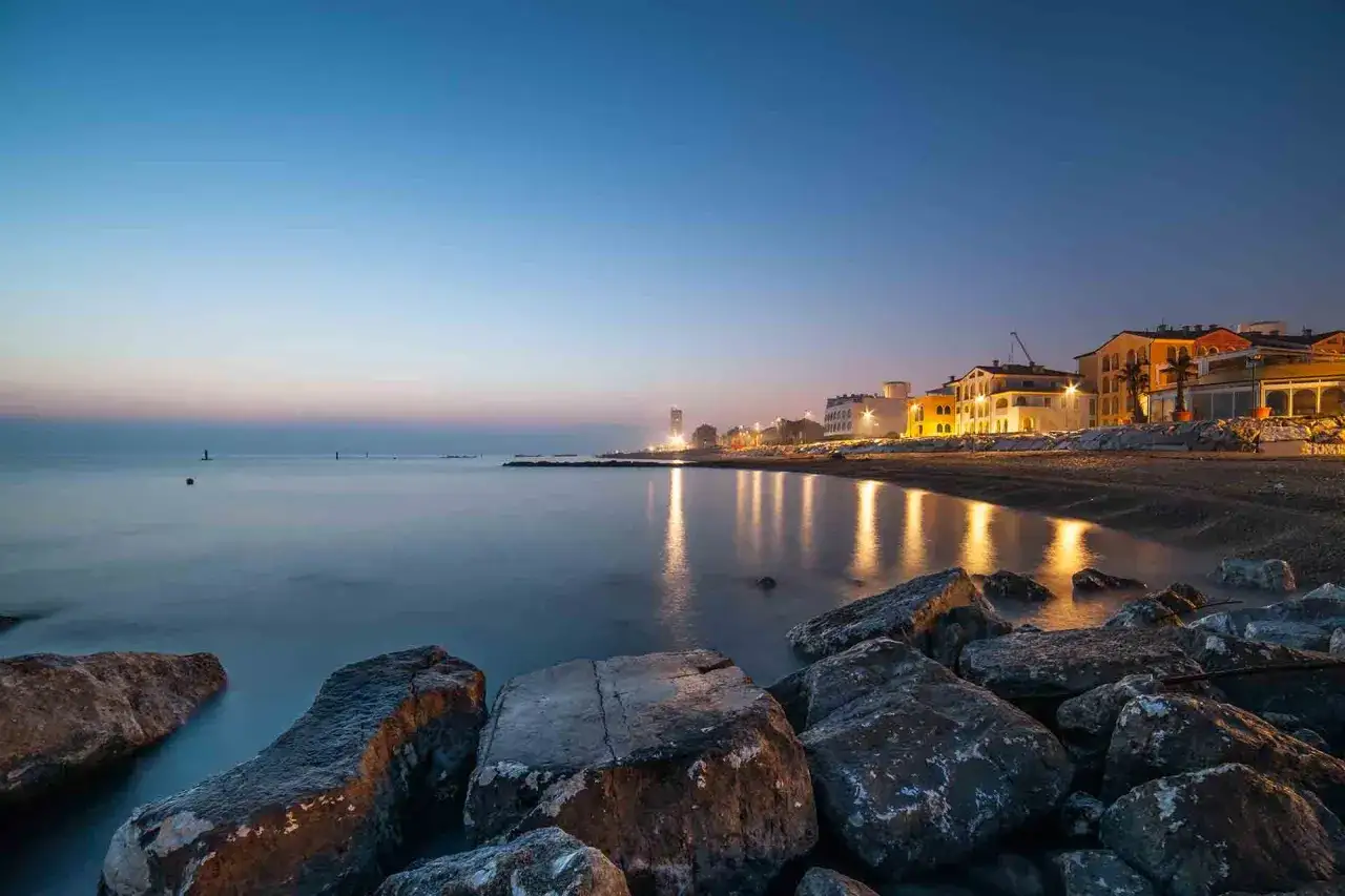 Spiaggia di Porto Recanati al crepuscolo, con luci che si riflettono sul mare calmo e rocce in primo piano. Un'atmosfera magica per scoprire cosa vedere a Porto Recanati.