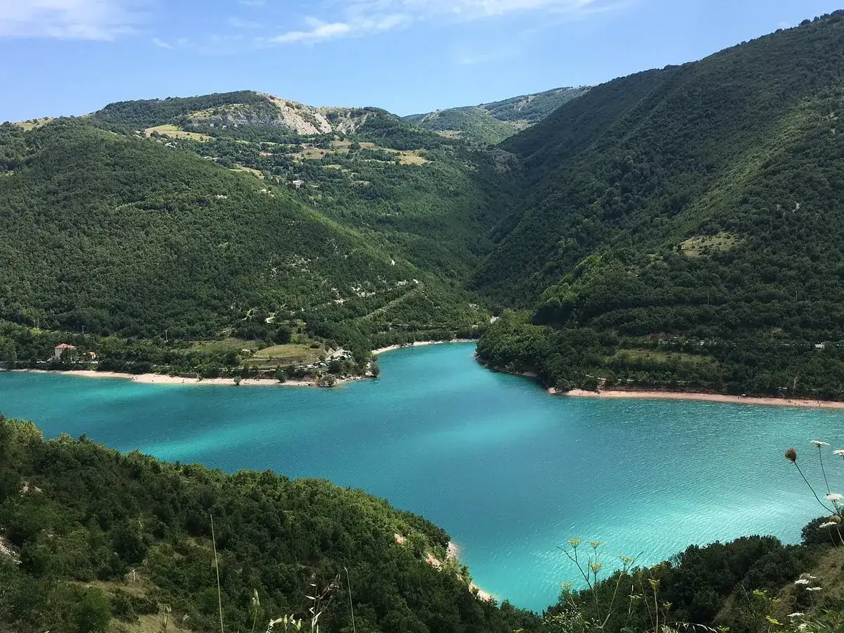 Splendida vista del lago di Fiastra con la sua spiaggia libera, circondato da verdi colline boscose sotto un cielo azzurro.