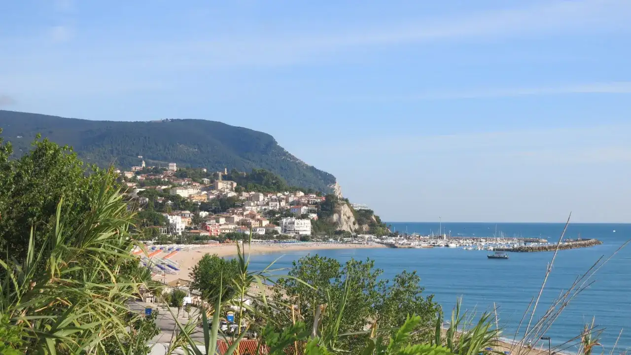 Vista panoramica del Monte Conero e del suo borgo affacciato sul mare Adriatico. Spiaggia attrezzata, porto turistico e vegetazione rigogliosa.