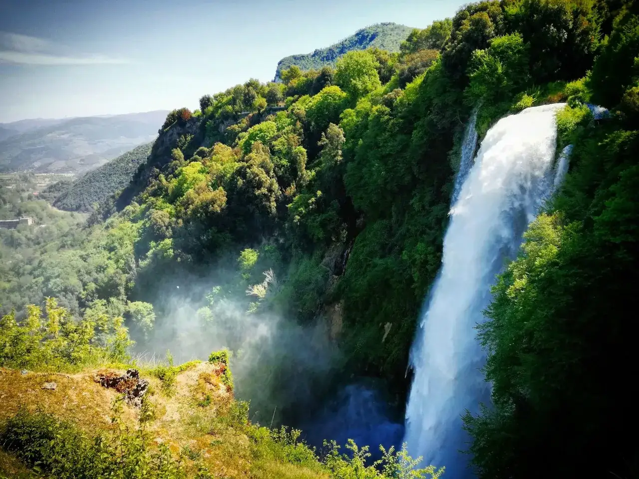Cascate delle Marmore immerse nel verde, con un sentiero che porta ai borghi vicini. Nebbia e acqua creano un'atmosfera magica.