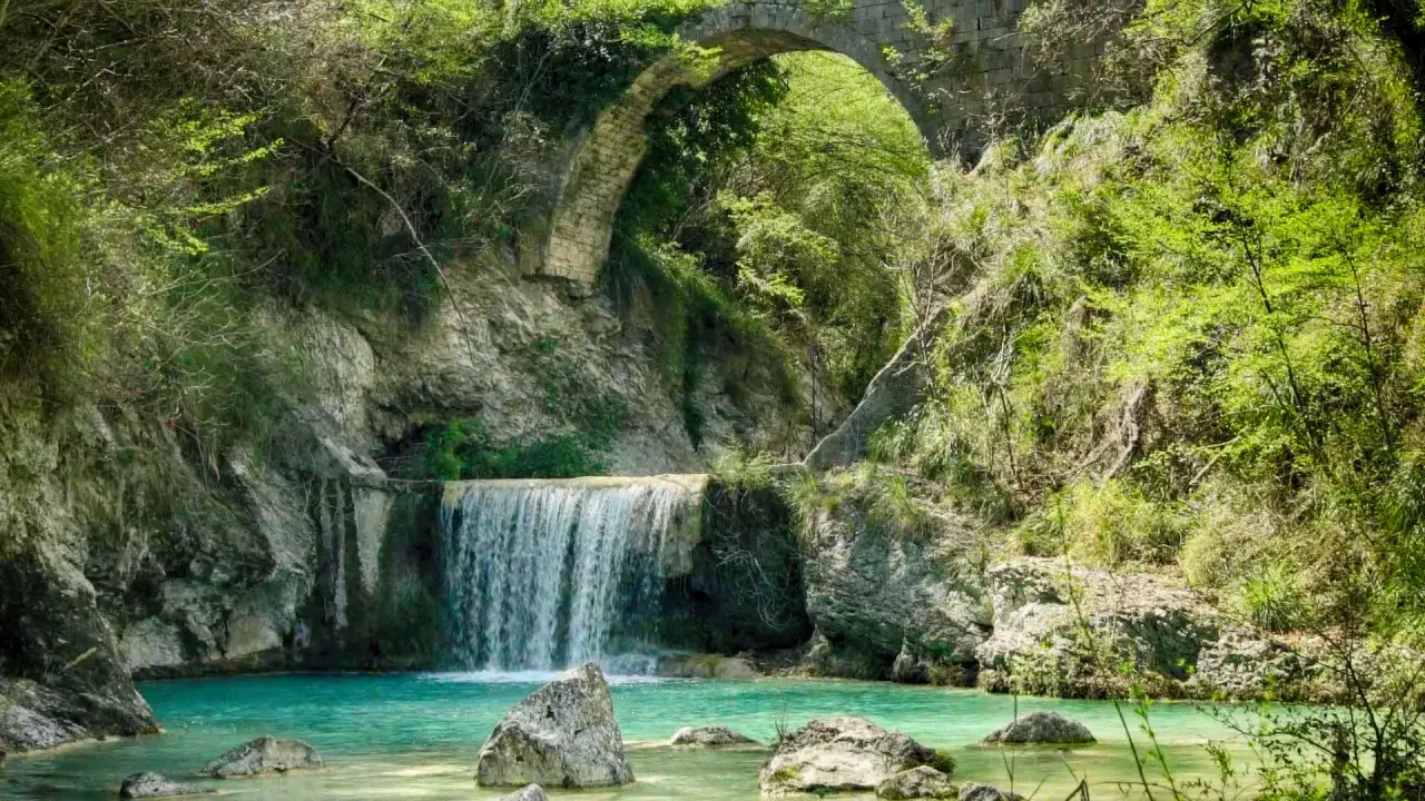 Cascata con acqua turchese sotto un antico ponte di pietra, immersa nel verde rigoglioso. Le cascatelle di Castel Trosino creano uno scenario incantevole.