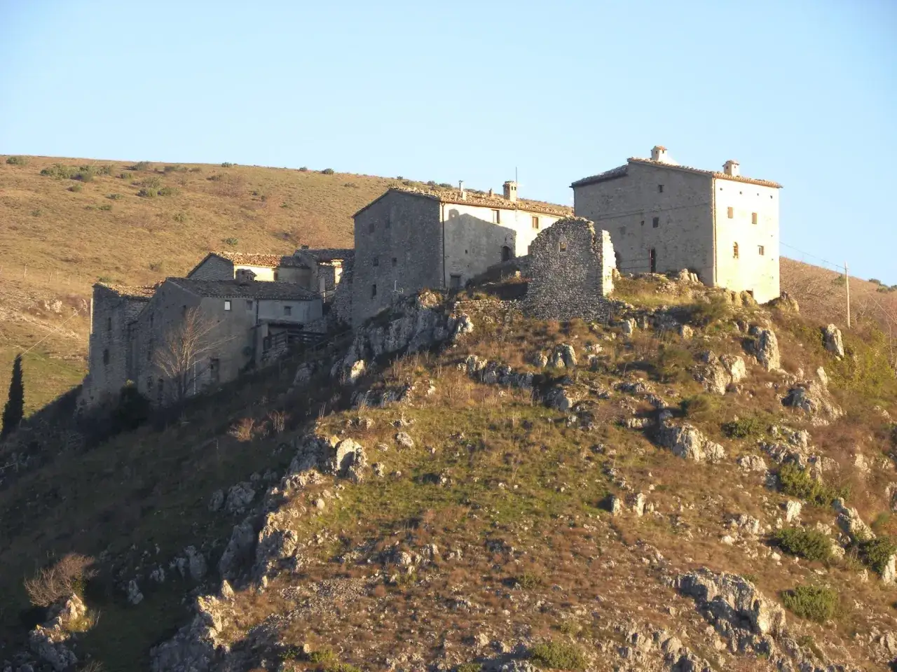 Antico borgo in pietra sui monti, un elcito nei Marche, con case arroccate su un crinale roccioso sotto un cielo limpido.