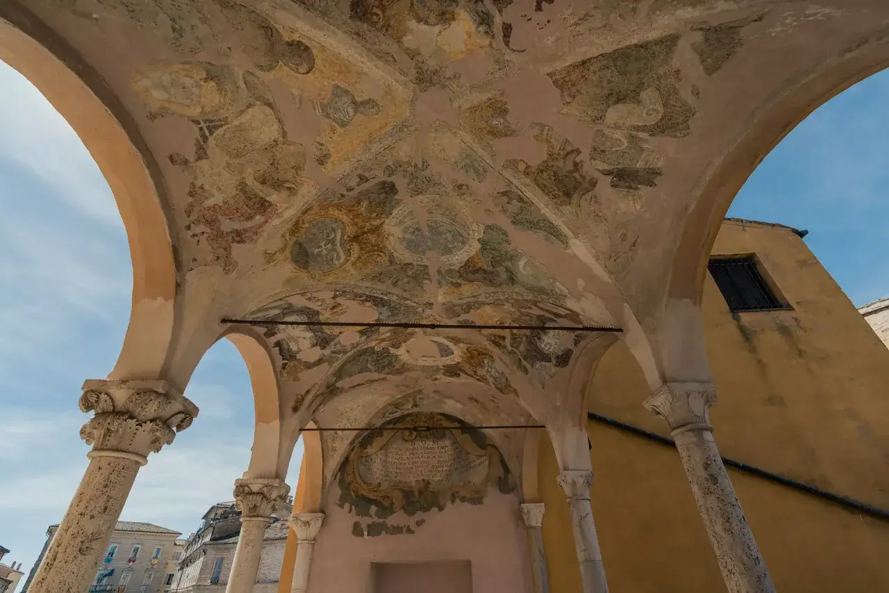 Soffitto affrescato di un portico a Fermo, nelle Marche. Colonne in pietra sorreggono archi decorati con scene antiche e stemmi.