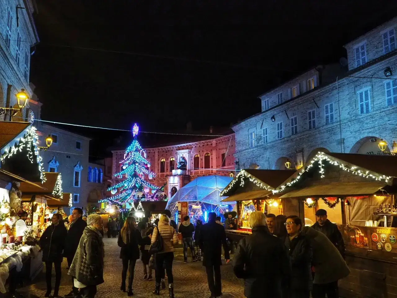 Atmosfera magica nei mercatini di Natale nelle Marche: gente passeggia tra bancarelle illuminate e un grande albero scintillante.