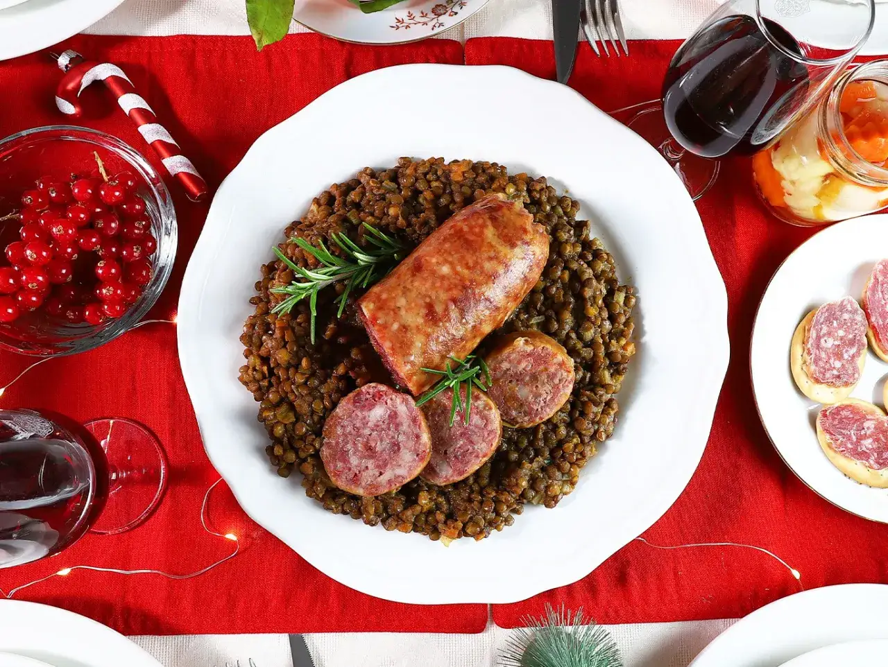 Piatto di lenticchie e cotechino alla marchigiana, guarnito con rosmarino, su un tavolo natalizio con ribes, vino rosso e salumi.