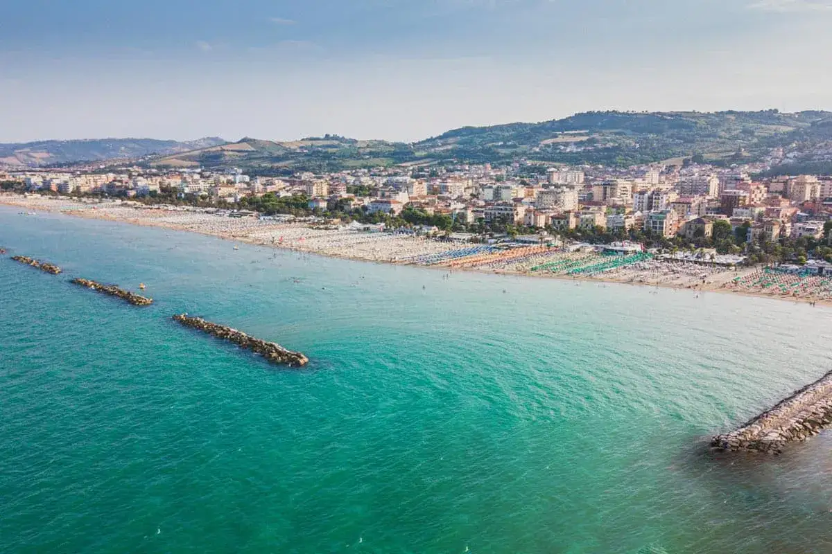 San Benedetto del Tronto: un'ampia spiaggia di sabbia dorata con file di ombrelloni colorati e un mare pulito e cristallino. La citt&agrave; si estende sullo sfondo, con colline verdi che la abbracciano.