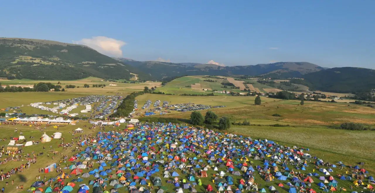 Vasto accampamento di tende colorate per il festival celtico Montelago, con montagne sullo sfondo e un parcheggio pieno di auto.