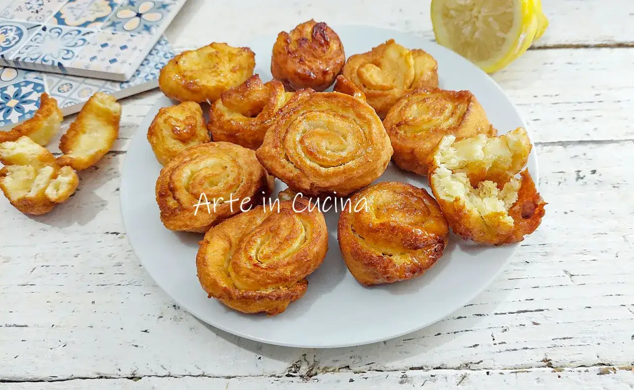 Piatto di dolci fritti, alcuni a spirale, altri a forma di fiore, che ricordano i limoncini dolci. Un limone a fette sullo sfondo.