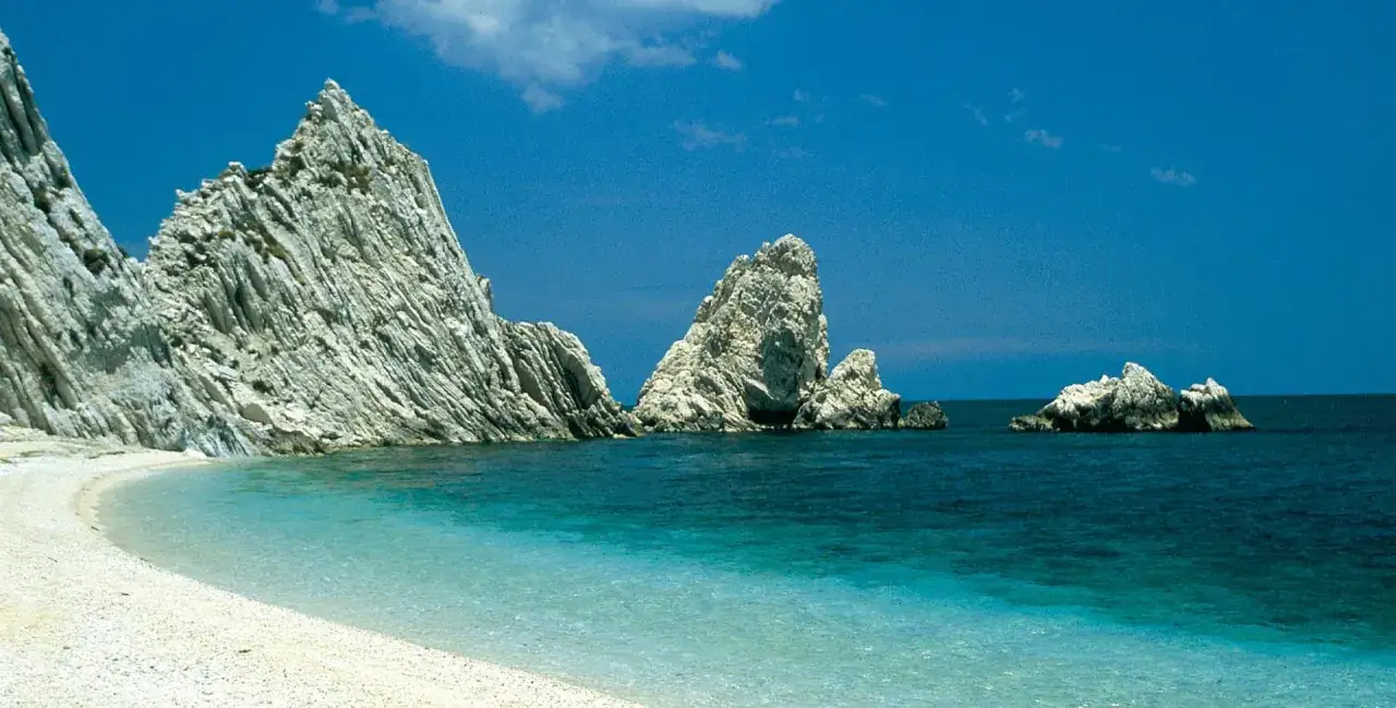 Spiaggia bianca e mare cristallino sulla riviera marchigiana, con imponenti faraglioni che emergono dall'acqua blu intenso sotto un cielo sereno.