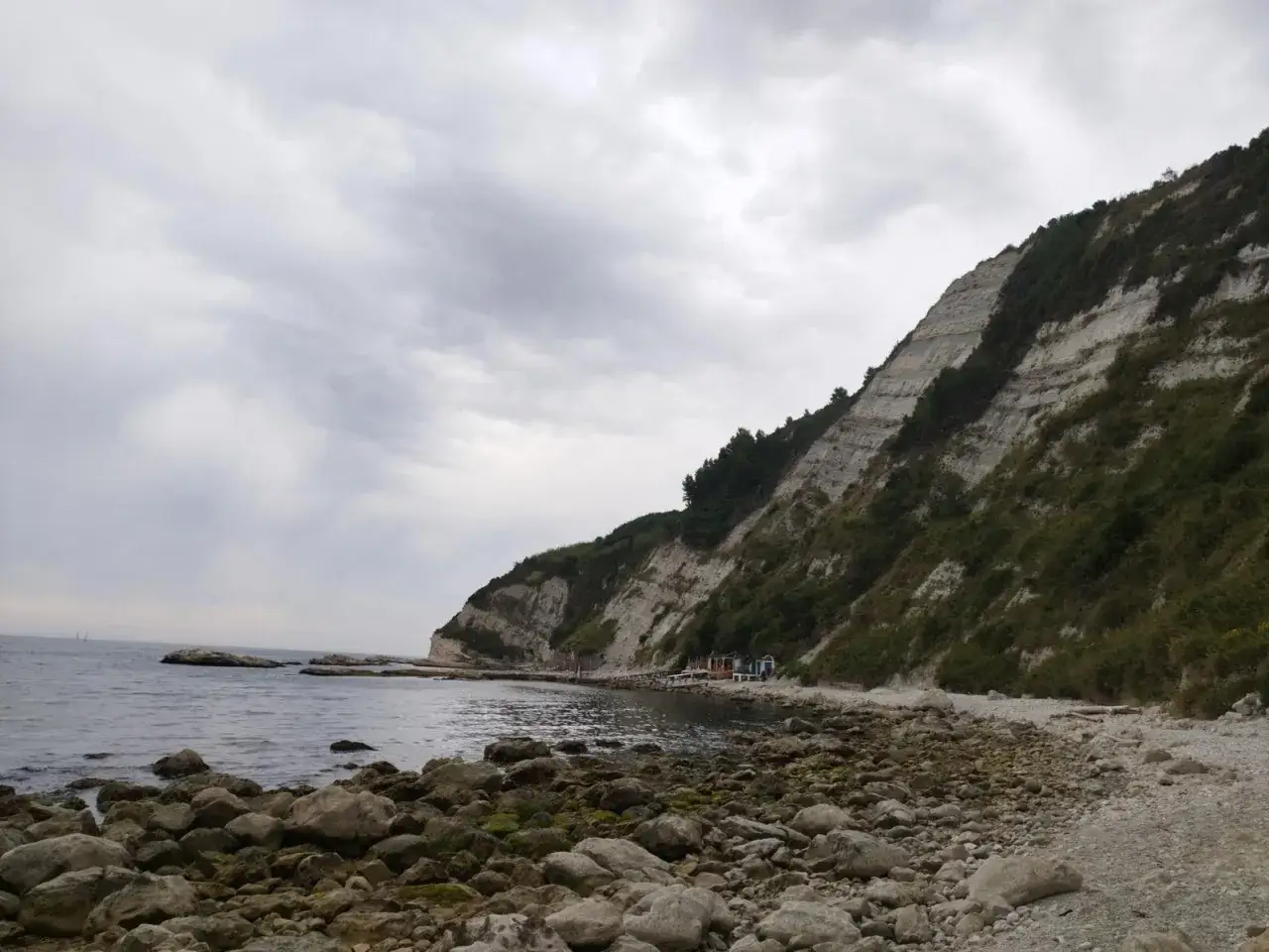 Scogliera verdeggiante e mare calmo alla spiaggia della Scalaccia, con rocce levigate sulla riva e un cielo nuvoloso.