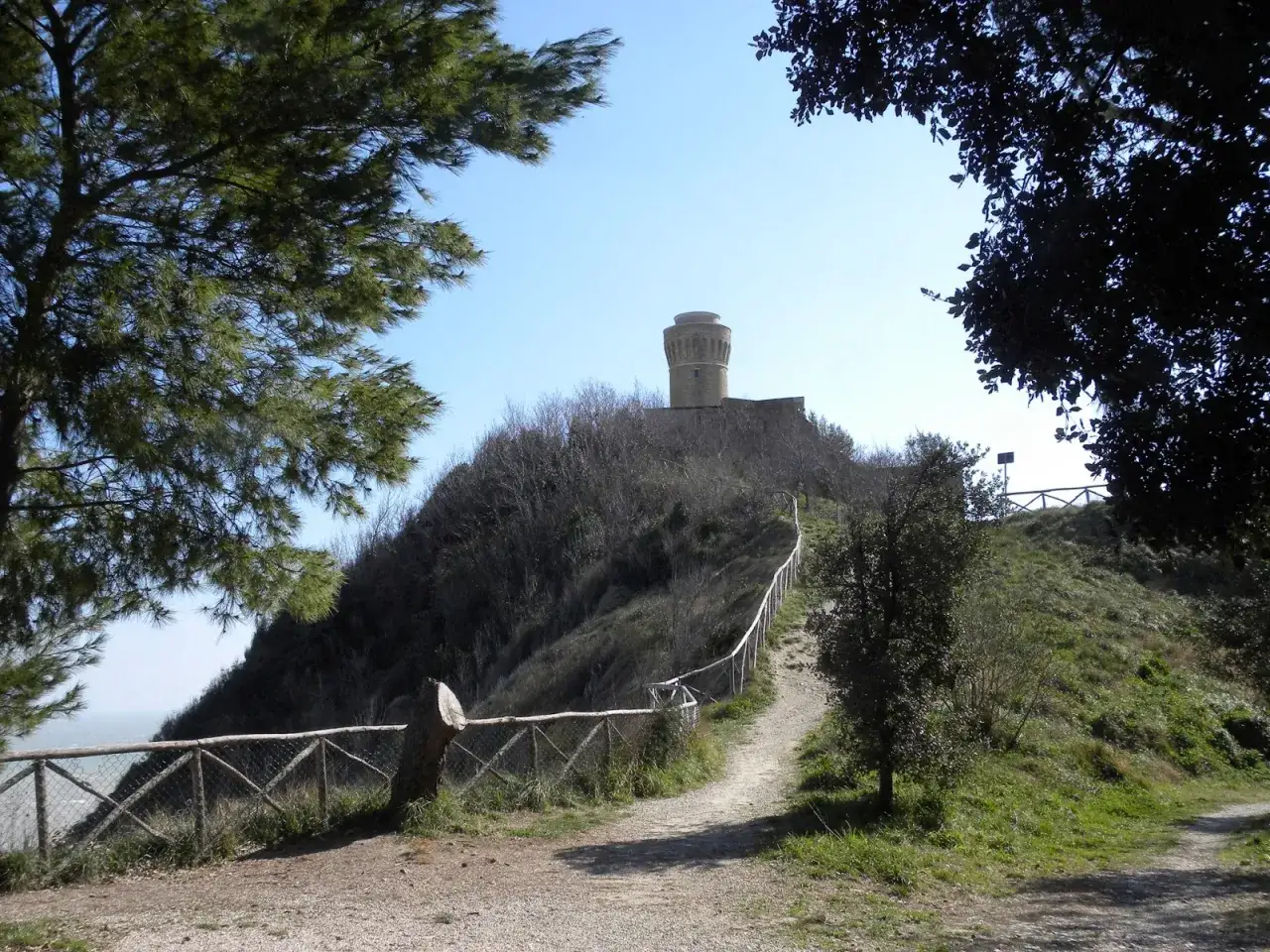 Sentiero nel parco del cardeto che sale verso una torre circolare su una collina brulla, con alberi e recinzione in legno.