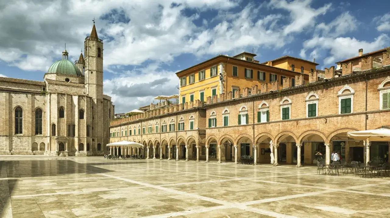 Piazza del Popolo ad Ascoli Piceno: cosa vedere. Maestosa cattedrale con cupola verde, portici eleganti e palazzi storici creano uno scenario unico.