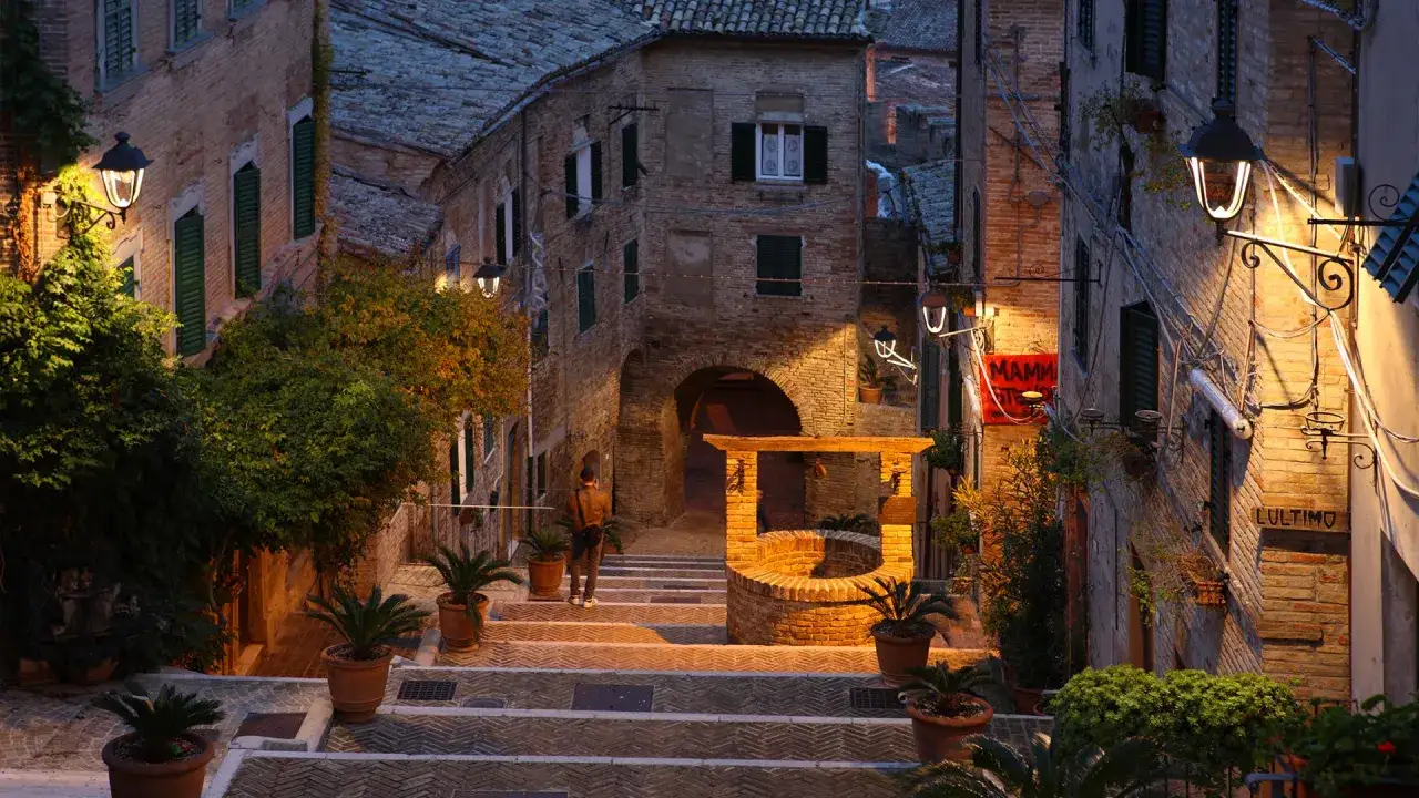 Serra de' Conti: scalinata illuminata da lampioni, un pozzo antico e un arco in pietra creano un'atmosfera magica al tramonto.
