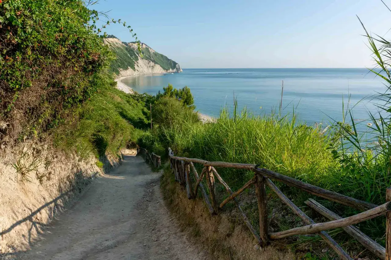 Sentiero sabbioso in mezzavalle discesa, con staccionata in legno e vista sul mare Adriatico e le scogliere.