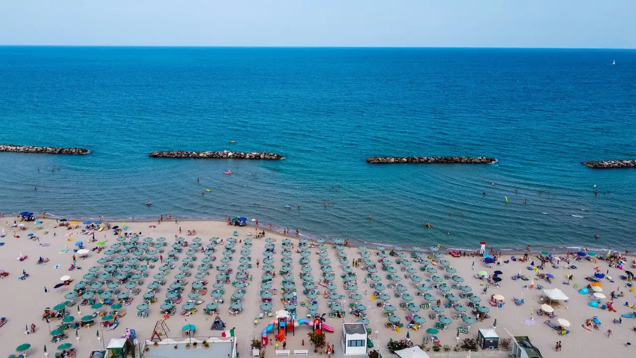 Spiaggia affollata con ombrelloni verdi e blu, bagnanti in mare e frangiflutti in pietra. Un'immagine di un perfetto giorno di fermo mare.