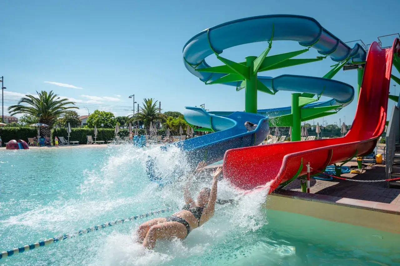 Divertimento assicurato in questo parco acquatico nelle Marche: una ragazza scivola tra spruzzi d'acqua su uno scivolo rosso e blu.