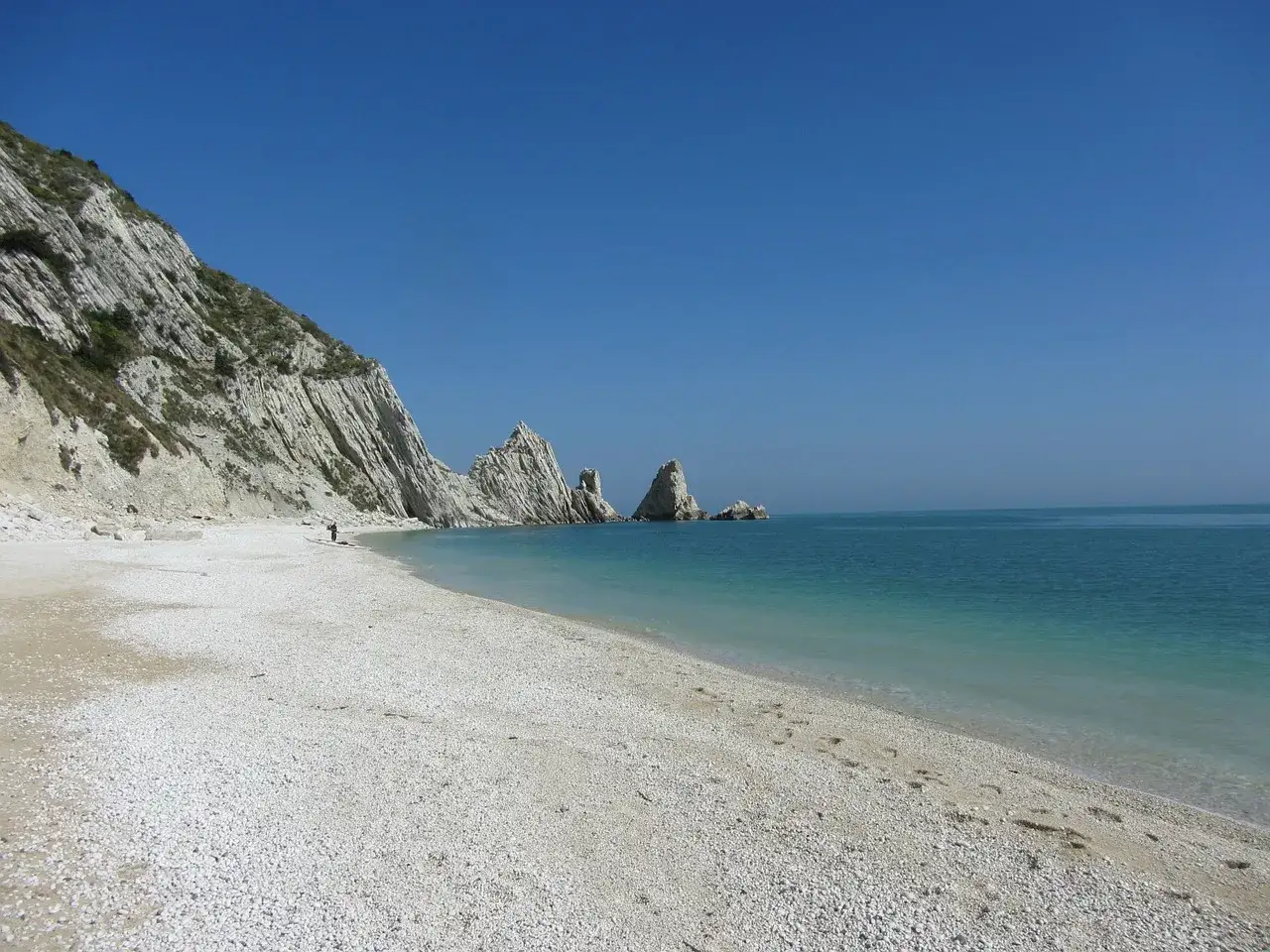 La spiaggia delle Due Sorelle, con le sue iconiche rocce, si estende sotto un cielo azzurro. L'acqua cristallina lambisce la riva di ciottoli bianchi.