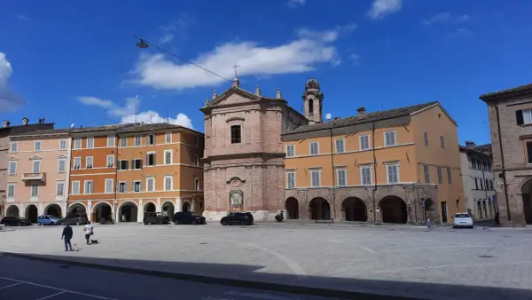 Piazza a San Severino Marche con edifici colorati, una chiesa in mattoni e cielo azzurro.
