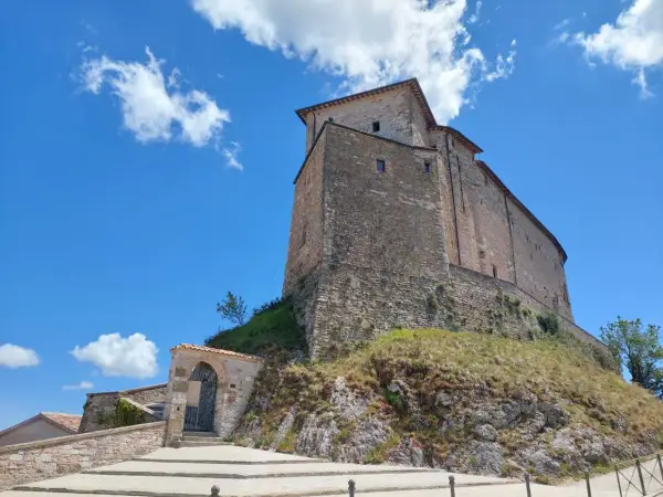 Antico borgo di Frontone, Marche, con imponente rocca in pietra su un colle erboso, sotto un cielo azzurro con nuvole bianche.